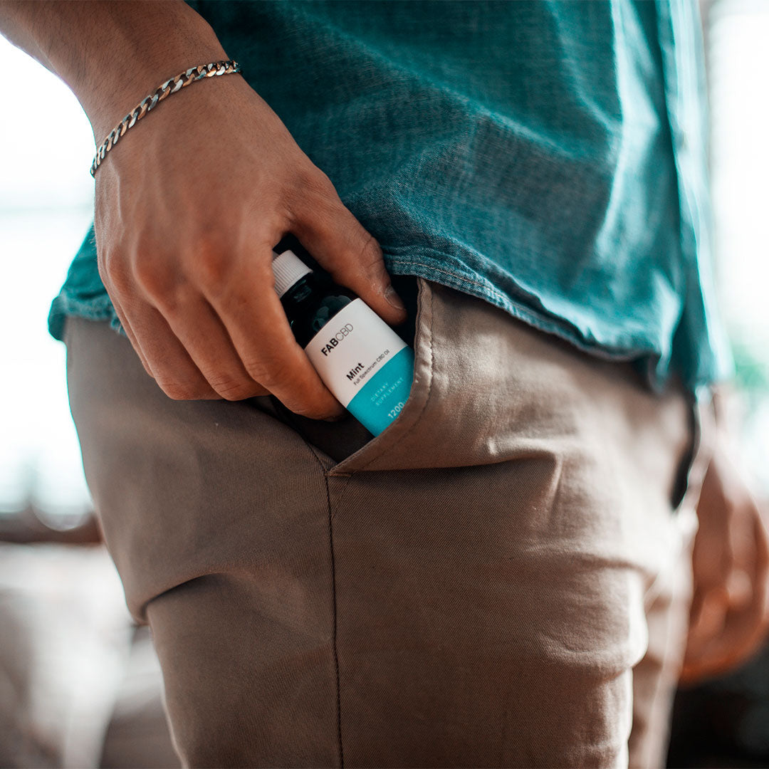 Close-up of a hand sliding a FAB CBD CBD Oil bottle into the front pocket of khaki pants against a teal shirt.