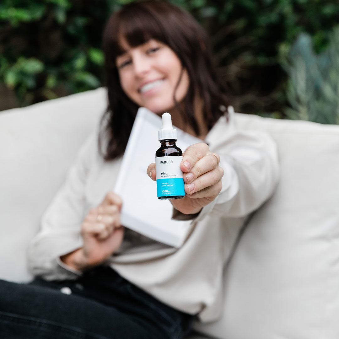 Woman reclining on a couch holding a small CBD Oil dropper bottle toward the camera, label and teal accent clearly visible.