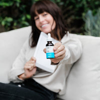 Woman reclining on a couch holding a small CBD Oil dropper bottle toward the camera, label and teal accent clearly visible.