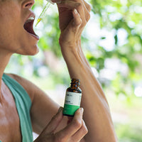 Close-up of a person tilting a dropper toward their open mouth while holding a small amber bottle labeled "CBD Oil" against a soft-focus green outdoor background