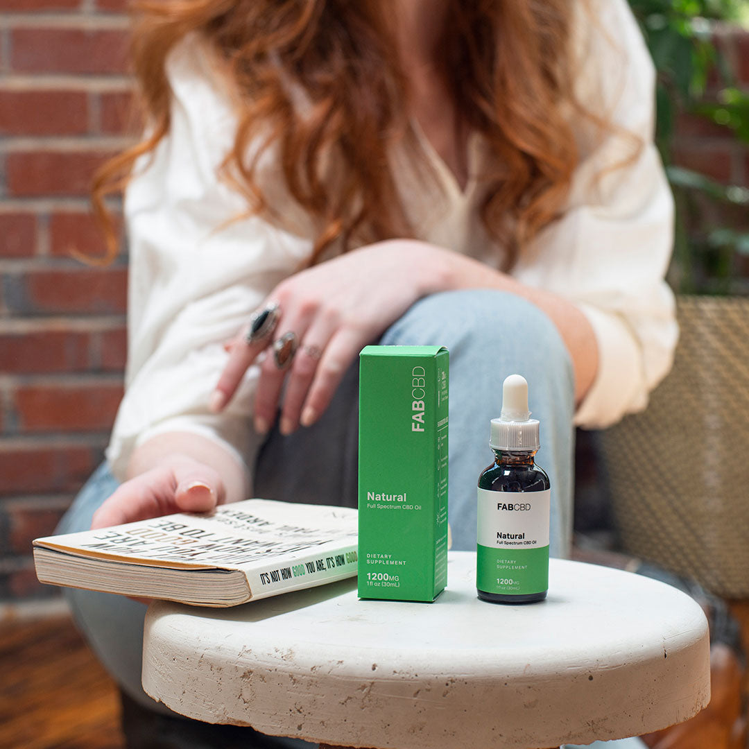 Three-quarter view of the FABCBD CBD Oil bottle and green box on a round white stool in front of a seated person reading a book.