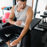 Side view of a man seated on a gym bench applying CBD Body Salve to his forearm, with weights and a red exercise ball in soft daylight.