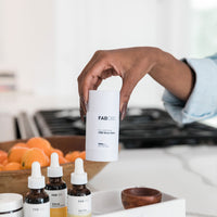 Close-up of a hand holding a white FAB CBD CBD Body Salve stick above a kitchen counter with dropper bottles and a bowl of oranges in soft natural light
