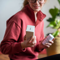 Close-up of a person holding a white box labeled FAB CBD CBD Body Salve, angled toward the camera with a blurred home plant background.