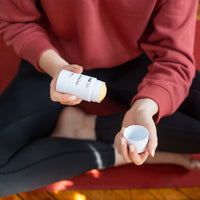 Close-up of hands unscrewing a white tube of CBD Body Salve to reveal the yellow balm, seated cross-legged on a red mat.