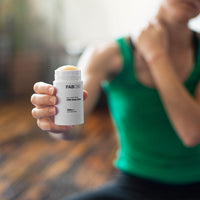 Close-up of a hand holding a white stick of FABCBD CBD Body Salve toward the camera with a blurred person in a green tank top applying it to their neck.