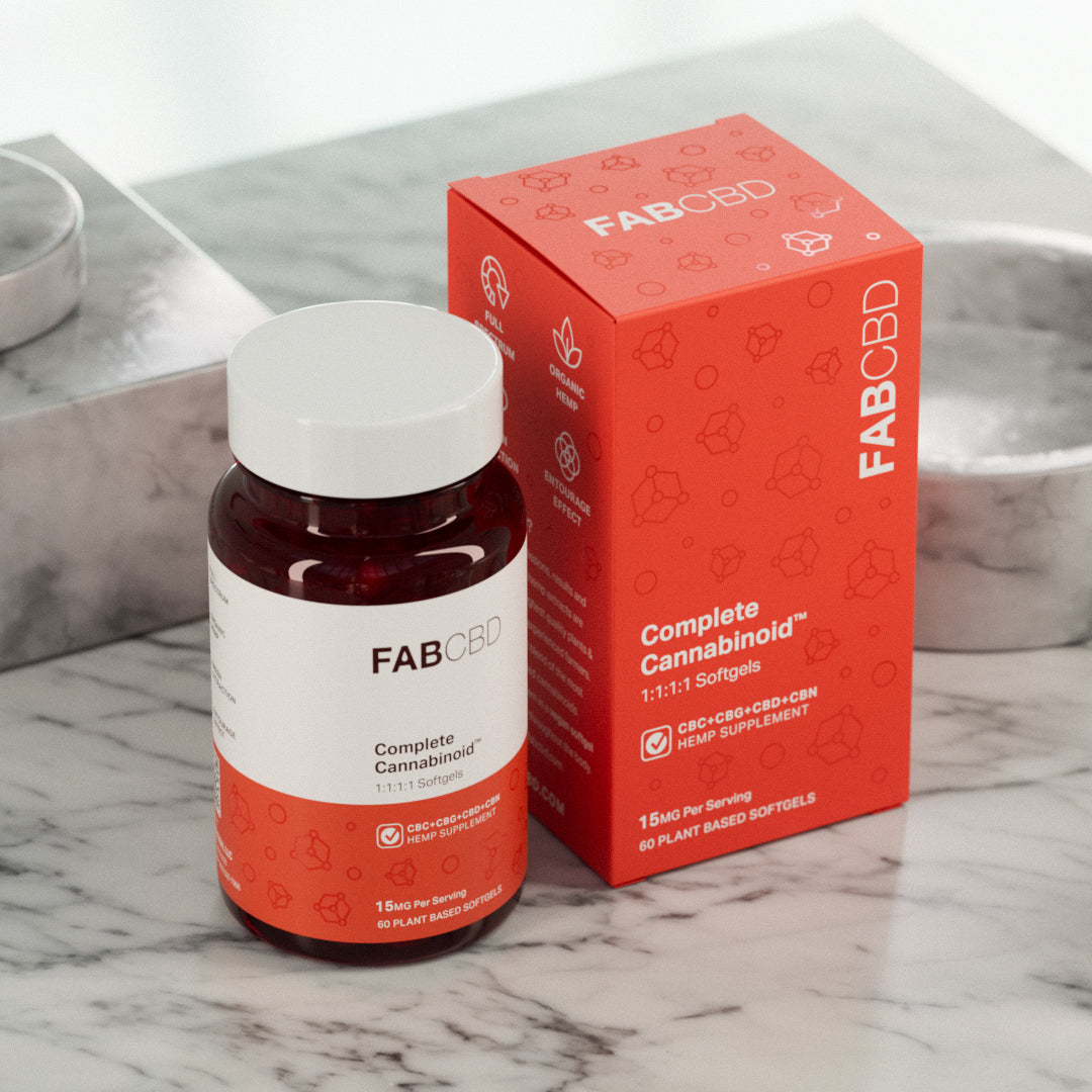 An upright amber glass supplement bottle with a white cap sitting next to its matching bright orange packaging box on a grey marbled surface. Both the bottle and the box showcase FAB CBD branding for Complete Cannabinoid™ 1:1:1:1 Softgels and explicitly display the product specifications: "CBC+CBG+CBD+CBN HEMP SUPPLEMENT," a potency of "15MG Per Serving," and a count of "60 PLANT BASED SOFTGELS."