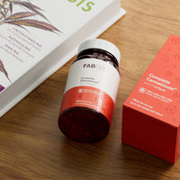 An amber glass supplement bottle with a white cap resting at an angle on a wooden surface next to its bright orange packaging box and a book. Both the bottle and the box feature FAB CBD branding for Complete Cannabinoid™ 1:1:1:1 Softgels and display key product specifications: "CBC+CBG+CBD+CBN HEMP SUPPLEMENT," a potency of "15MG Per Serving," and a count of "60 PLANT BASED SOFTGELS."