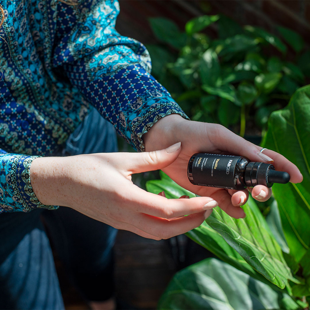 Close-up of hands holding a small dark glass dropper bottle labeled CBD Oil against leafy green plants and sunlight.