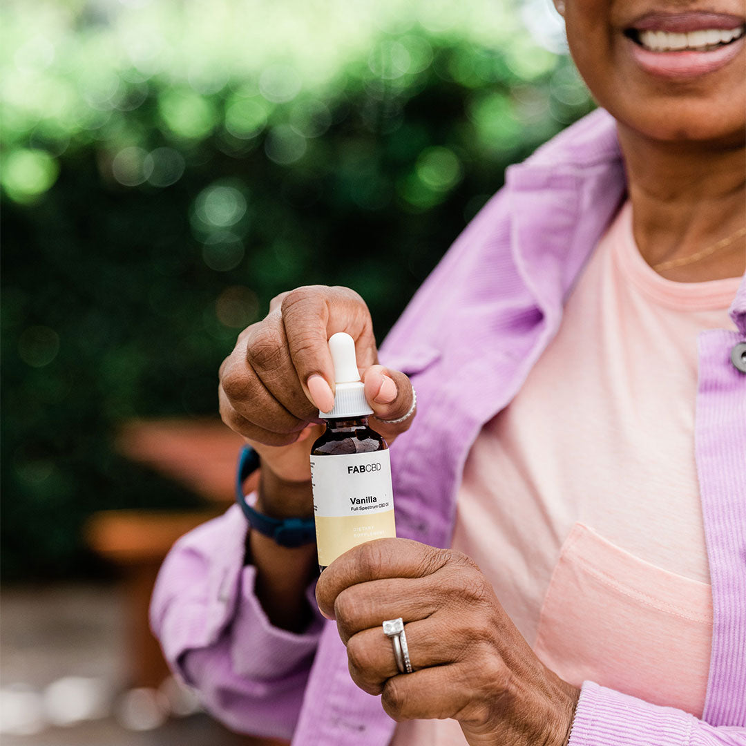 A close-up of a person holding a small amber dropper bottle labeled CBD Oil against a soft-focus green outdoor background.