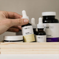 A hand reaching for a FABCBD CBD Oil dropper bottle labeled "Vanilla" among other FABCBD tinctures and jars in a wooden tray.