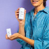 Woman holding two white sticks of FAB CBD CBD Body Salve against a purple backdrop, close-up of hands and torso in a blue shirt.